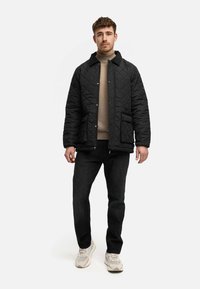 Zwarte gewatteerde jas met een kraag, vooraan met drukknopen, twee voorzakken, gedragen over een beige trui, gecombineerd met zwarte jeans en lichte sneakers.