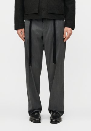 Άνδρας που φορά χαλαρά γκρι παντελόνια με ρίγες, μακριά μαύρα κορδόνια, μαύρο μπουφάν και μαύρα δερμάτινα παπούτσια, στέκεται ακίνητος.