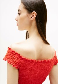 Top rosso off-the-shoulder in tessuto strutturato con design arricciato; orli frastagliati e silhouette aderente mettono in risalto il colletto e le spalle.