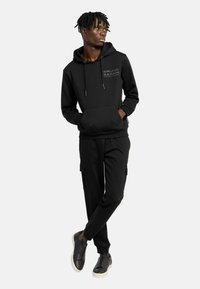 Zwarte hoodie met koordjes en een voorzak, gecombineerd met bijpassende joggers met cargozakken. Draagt met zwarte sneakers.