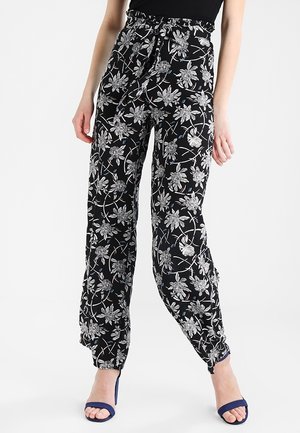 Vrouw draagt zwarte, hoog getailleerde broek met witte bloemenprint en blauwe sandalen met hak, staand tegen een witte achtergrond.