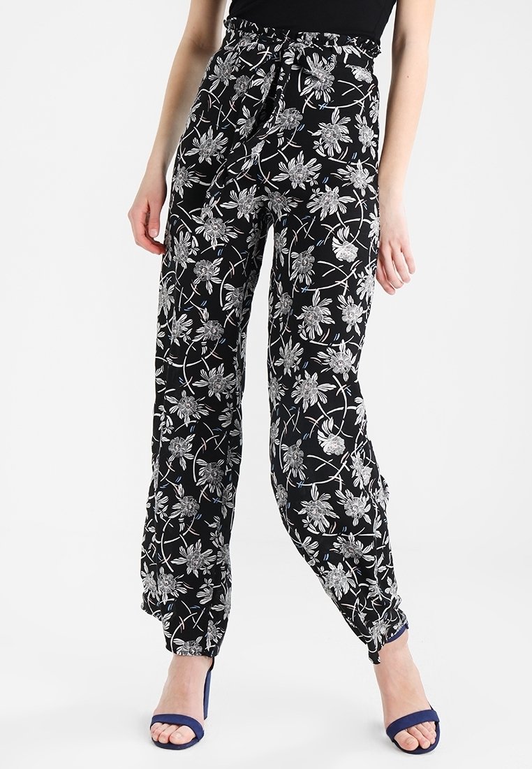Femme portant un pantalon noir taille haute à imprimé floral blanc et des sandales bleues à talons, debout devant un fond blanc.