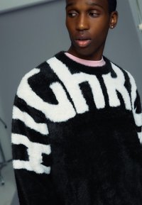 Pull flou noir et blanc avec un lettrage audacieux "HIGH", présentant un col rond et une chemise rose superposée en dessous. Texture lisse.