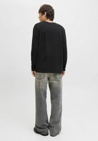 Persona con cabello corto oscuro vestida con una camiseta negra de manga larga y unos pantalones anchos de mezclilla gris descolorido, de pie dando la espalda sobre un fondo blanco liso.