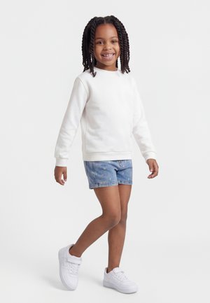 Lächendes Mädchen mit geflochtenem Haar, das einen weißen Pullover, Jeansshorts mit kleinen farbigen Sternen und weiße Turnschuhe trägt und auf einem Bein steht.