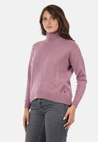 Donna che indossa un maglione a collo alto mauve e jeans grigi, in piedi con una mano sui fianchi contro uno sfondo bianco.