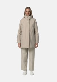 Beżowa kurtka wierzchnia z wysokim kołnierzem i zamkiem z przodu, z bocznymi kieszeniami i minimalistycznym designem. Noszona z dopasowanymi spodniami i białymi trampkami.