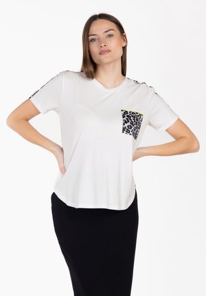 Jeune femme aux cheveux bruns raides portant un t-shirt blanc avec une poche imprimée léopard et des rayures, associé à une jupe noire, mains sur les hanches.