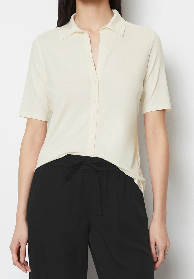 Camisa de manga corta en un tono beige claro con cuello, cierre frontal de botones y textura suave, combinada con pantalones negros con cordón.