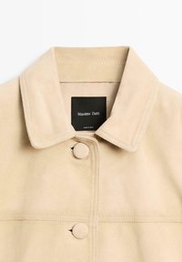 Beige ruskindsjakke med klassisk krave, knaplukning og diskrete syningsdetaljer. Har et sort mærke med brandnavnet indeni.