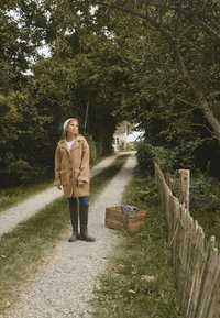 Béžový kabát s ovčím golierom a manžetami, modré džínsy a zelené čižmy po kolená; stojí na štrkovom chodníku lemovanom zeleňou.