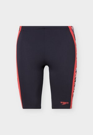 Ναυτικό μπλε ανδρικό μαγιό Speedo τύπου jammers με κόκκινες ρίγες στο πλάι και σχεδιασμένες επιφάνειες στα εξωτερικά μέρη, εφαρμοστή γραμμή μέχρι το γόνατο.