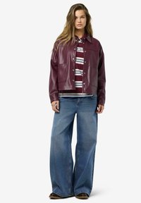Burgunderfarbene Kunstlederjacke mit Druckknöpfen, kombiniert mit einem gestreiften, kastanienbraunen und weißen Hemd sowie weit geschnittenen blauen Jeans.