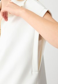 Witte mouwloze top met een ritssluiting aan de voorkant, zachte textuur en geplooide schoudernaad, die een binnenzak aan de zijkant onthult.