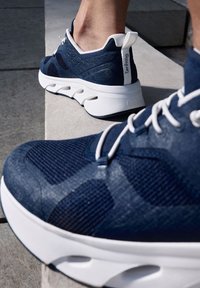 Baskets de sport bleu marine en tissu mesh, avec des lacets blancs et une semelle blanche rembourrée. Elles présentent des accents texturés et une bride à l'arrière.