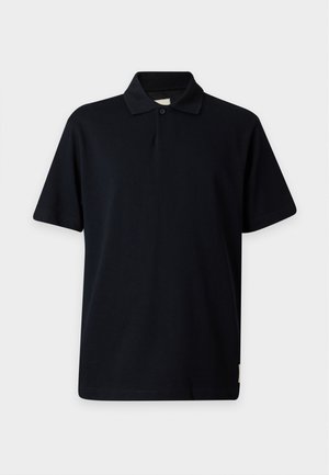 Jeune homme debout portant un polo noir, un jean gris et des baskets noires avec des semelles blanches sur un fond blanc uni.