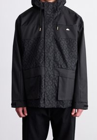 Svart jacka med ett strukturerat mönster på framsidan, justerbar huva, framfickor och en slät yta. Guldiga detaljer är inkluderade.