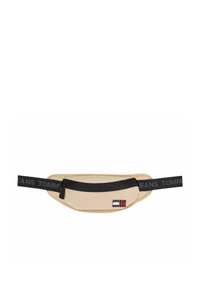 Riñonera beige fabricada en tela, con una correa ajustable negra etiquetada con "TOMMY JEANS" y un pequeño parche de logo con elementos en rojo, blanco y azul.