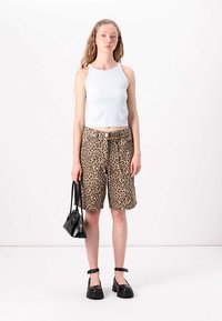 Biely rebrovaný top, hnedé šortky s leopardím vzorom, čierne topánky na členkovom popruhu a malá čierna kabelka. Modelka stojí pred jednoduchým pozadím.