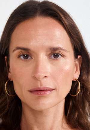 Žena smeđe kose i smeđih očiju, nosi zlatne naušnice u obliku obruča, gleda naprijed s neutralnim izrazom lica na jednostavnoj pozadini.