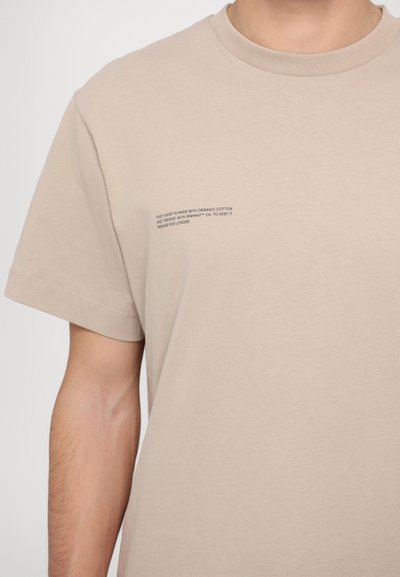 Tricou bej din bumbac organic, cu mâneci scurte și decolteu rotund. Text mic pe piept care detaliază tratamentul materialului.