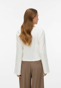 Hvid bluse med lange ærmer og rund hals, kombineret med brune højtaljede bukser med to baglommer og knapdetaljer.