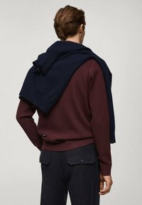Pull côtelé marron avec un châle en tricot bleu marine drapé sur les épaules, accompagné d'un pantalon bleu marine avec des poches arrière.