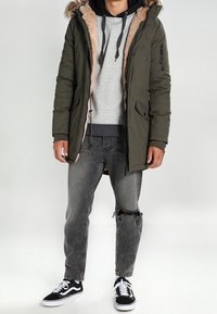Parka verde oliva con fodera in pelliccia sintetica, chiusura con zip e tasche laterali. Felpa con cappuccio grigia sotto, abbinata a jeans grigi strappati e sneakers nere.