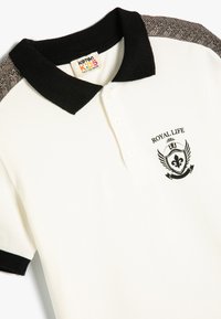 Hvid polo shirt med en sort strikket krave og ærmer. Har et broderet "Royal Life" logo på brystet og strukturerede skulderdetaljer.
