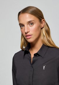 Chemise noire à col, en tissu lisse, avec un petit motif brodé blanc sur la poitrine gauche et une fermeture par bouton au niveau du cou.