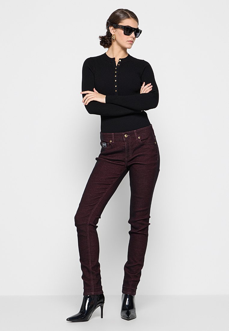 Czarny prążkowany top z długim rękawem z złotymi guzikami, w połączeniu z bordowymi rurkami o subtelnym połysku i złotymi detalami, noszone z czarnymi szpilkami.