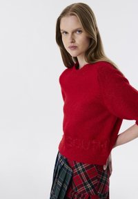 Roter gestrickter Pullover mit kurzen Ärmeln, der eine strukturierte Oberfläche aufweist und das Wort "SOUTH" am unteren Rand geprägt hat, kombiniert mit einem karierten Rock.