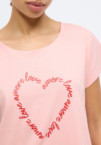Ροζ ανοιχτόχρωμο T-shirt με στρογγυλή λαιμόκοψη, με κόκκινη καρδιά σχεδιασμένη από τη λέξη "αγάπη" σε καλλιγραφική γ ραμματοσειρά. Κοντά μανίκια.