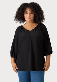 Blusa nera con scollo a V e dettagli arricciati sulle spalle, maniche puff a 3/4. Tessuto morbido con vestibilità rilassata, abbinata a denim blu.