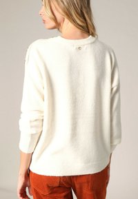 Pull en tricot crème, texture douce, larges manches trois-quarts, col rond, petit détail de bouton à l'arrière, associé à un pantalon en velours côtelé orange.