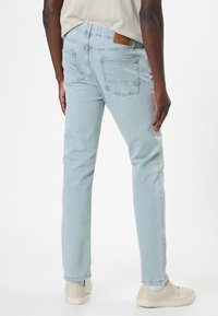 Vaqueros de mezclilla azul claro con un corte recto, dos bolsillos traseros y un parche de cuero marrón en la cinturilla, combinados con zapatillas blancas.