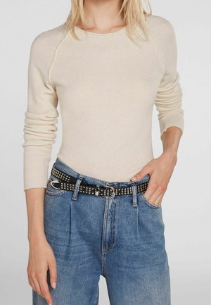 Femme portant un pull beige à manches longues, rentré dans un jean bleu taille haute avec une ceinture noire cloutée.