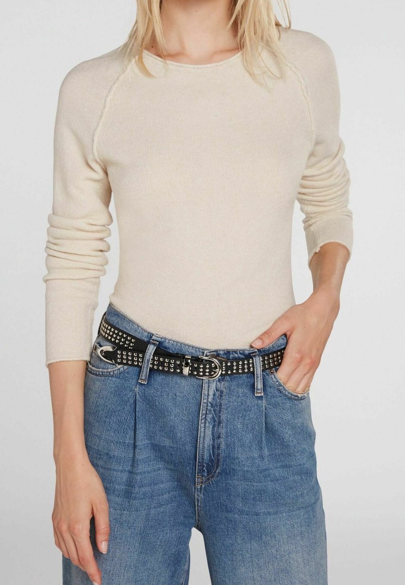 Femme portant un pull beige à manches longues, rentré dans un jean bleu taille haute avec une ceinture noire cloutée.
