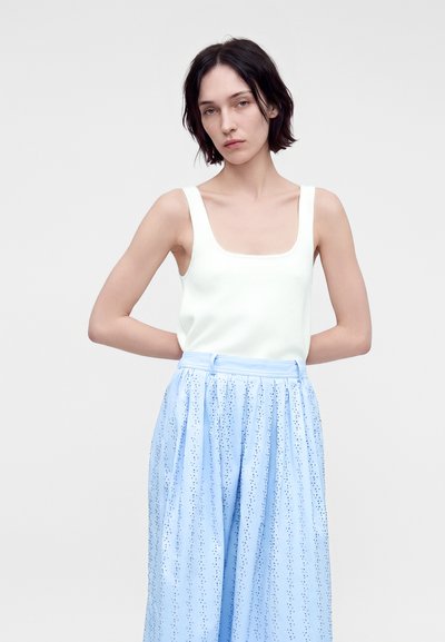 Jeune femme aux cheveux courts et foncés portant un haut blanc sans manches et une jupe plissée bleu clair taille haute avec de petits motifs à pois.