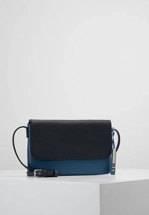 Kompakt crossbody taske med sort tekstureret klap, blå bund, justerbar rem og metal logo vedhæng på hvid overflade.