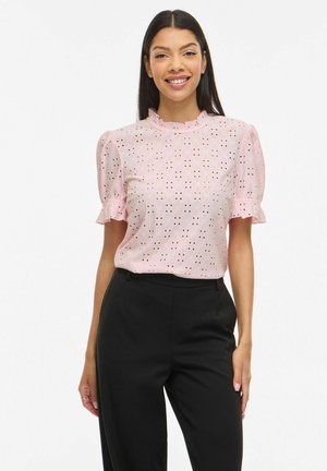 Femme portant un chemisier rose clair à œillets avec des manches courtes bouffantes et un pantalon noir taille haute, souriant à la caméra.