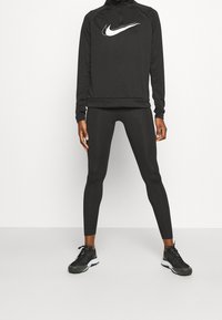Mustapohjainen urheilu pusero korkealla kaulalla ja Nike-logo, yhdistettynä mustiin leggings -housuihin ja mustiin urheilukenkiin, luoden virtaviivaisen siluetin.