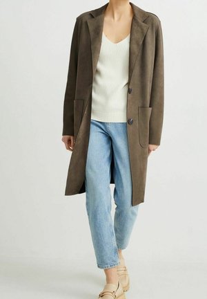 Personne portant un long manteau marron, un pull en maille crème, un jean bleu clair et des mocassins beige sur un fond clair uni.