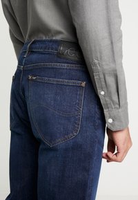 Ciemnoniebieskie dżinsy z kontrastującymi szwami, z czarną skórzaną naszywką oznaczoną napisem "Lee" w pasie oraz dwiema tylnymi kieszeniami.