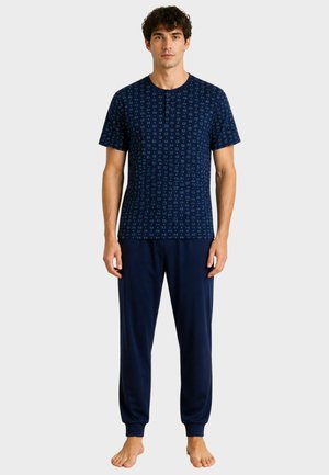 Uomo in piedi a piedi nudi che indossa una camicia blu navy a maniche corte con motivi e pantaloni da jogging blu navy tinta unita su sfondo semplice.
