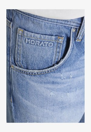 Primer plano del bolsillo de unos vaqueros de mezclilla azul con costuras amarillas y el nombre de la marca "MORATO" bordado en el pequeño bolsillo frontal.