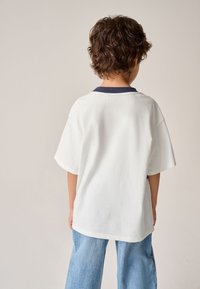 Fehér pamutpóló tengerészkék gallérral, rövid ujjakkal és laza szabással, világoskék farmernadrággal párosítva. Csak a hátsó nézet látható.