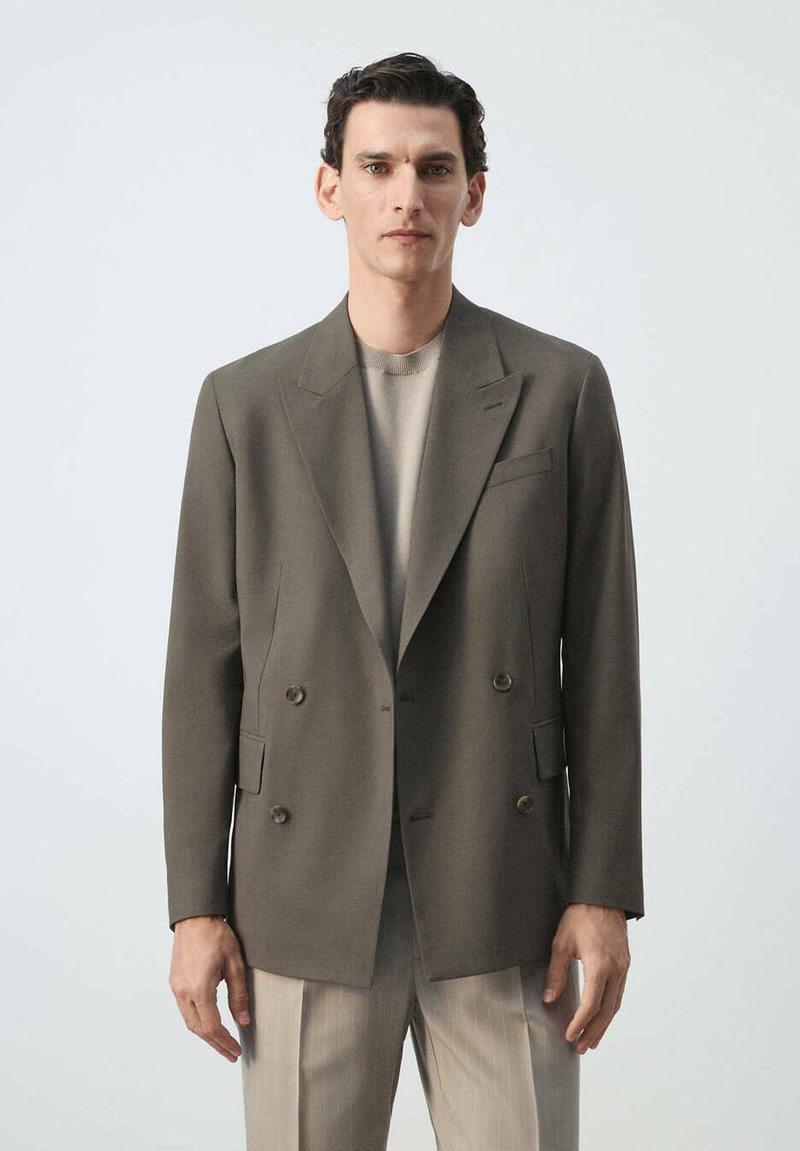 Homme aux cheveux courts et foncés portant un blazer croisé olive sur un pull beige et un pantalon beige clair, debout devant un fond uni.