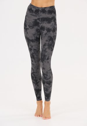 Mallas ajustadas de cintura alta con un patrón tie-dye en negro y gris oscuro, usadas descalzas, desde la cintura hasta los pies visibles sobre un fondo liso.