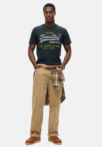 Donkerblauw katoenen t-shirt met vintage print, kaki katoenen broek en een geruit flanellen overhemd gestrikt rond de taille. Bruine veterlaarzen.